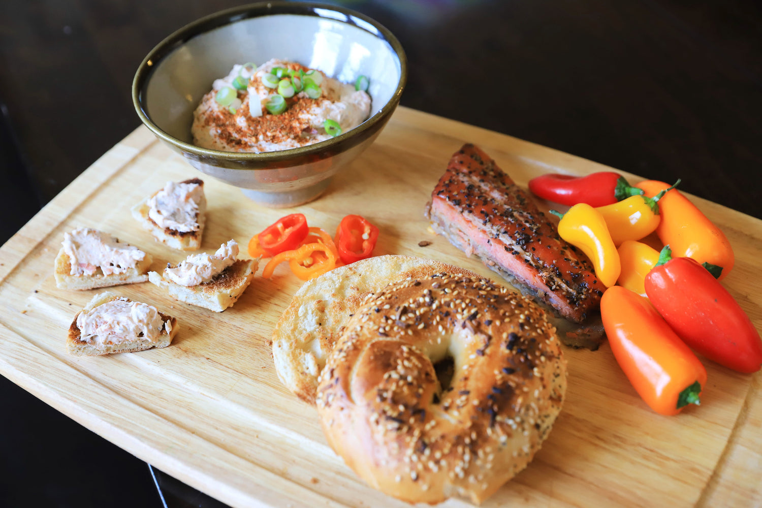 Wooden cutting board with bagel, cajun cream chesse, and colorful peppers on a dark background
